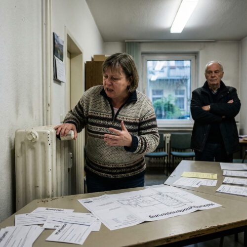 Die Hand einer Eigentümerin ruht besitzergreifend auf ihrem alten Rippenheizkörper während einer angespannten Versammlung.
