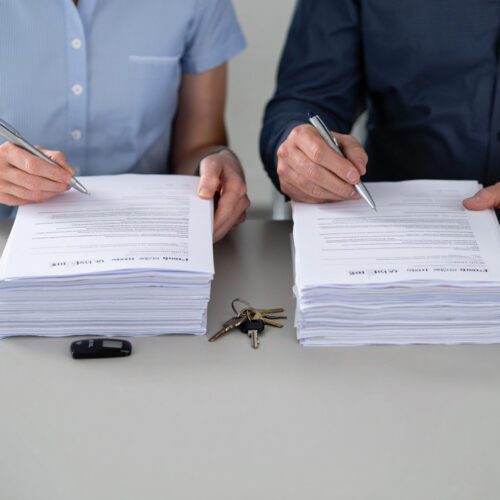 Zwei Personen unterzeichnen gleichzeitig separate, identische Vertragsstapel; ein Schlüsselbund und ein Transponder liegen dazwischen.