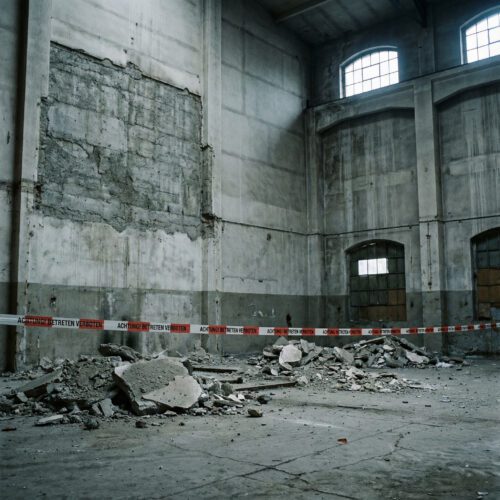 Massive Betonwand einer Industriehalle mit großen, abgebrochenen Putzplatten am Boden, die den rohen Untergrund freilegen.