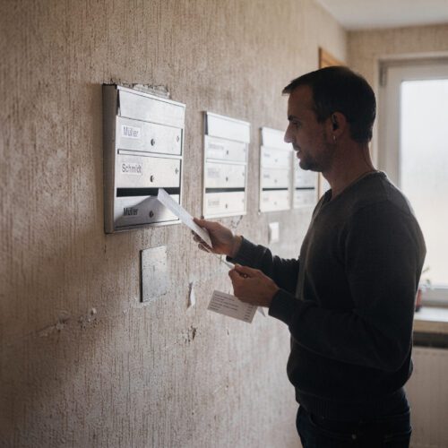 Eine Hand schiebt einen weißen Briefumschlag präzise in den metallischen Einwurfschlitz einer Briefkastenanlage.